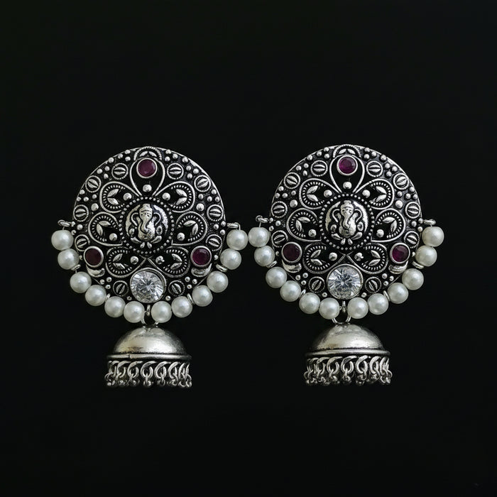 A pair of ornate silver-toned Indian jhumka earrings with intricate filigree work, white pearl bead borders, and small purple gemstone accents, displayed on a black stand against a dark background.