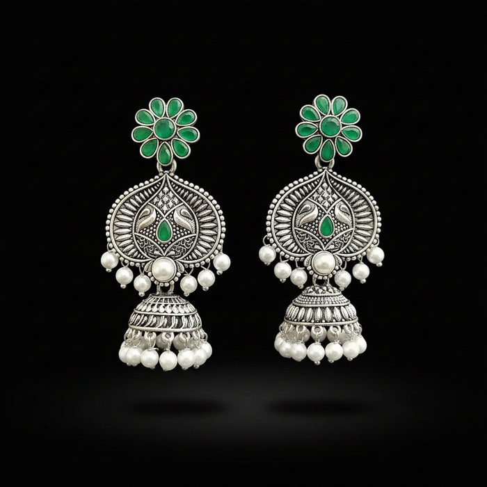 Ornate silver earrings with green gemstones and pearl-like beads on a gray stand.