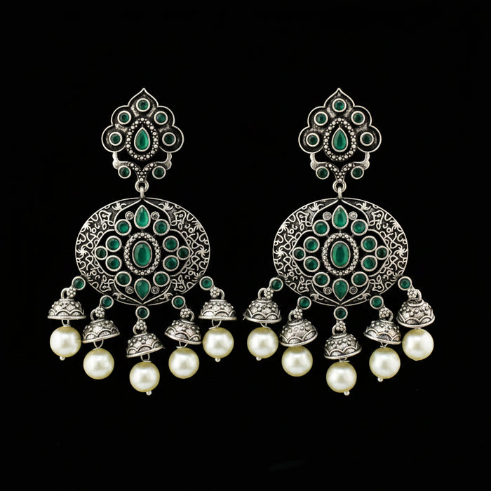 A pair of ornate silver-toned dangle earrings with green stone embellishments and white pearl drops, displayed on a gray stand against a gray background.