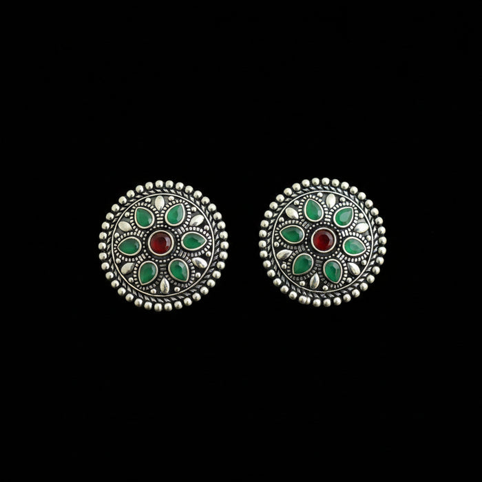 A pair of round silver-toned earrings with intricate filigree detailing, featuring green stone inlays and a central red stone, displayed on a dark reflective surface.