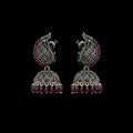 A pair of ornate silver-toned Indian jhumka earrings with peacock motifs, pink and green stone accents, and dangling bead chains, displayed on a black jewelry stand against a dark gray background.