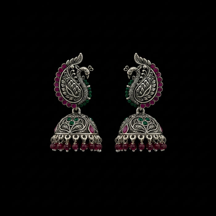 A pair of ornate silver-toned Indian jhumka earrings with peacock motifs, pink and green stone accents, and dangling bead chains, displayed on a black jewelry stand against a dark gray background.