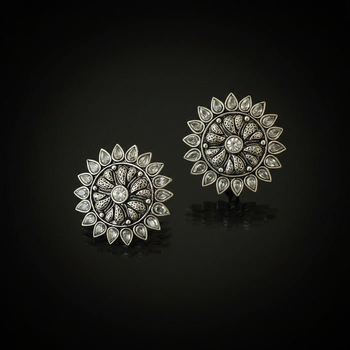 A pair of ornate silver-toned circular earrings with intricate filigree detailing and clear teardrop-shaped stones, displayed on a dark reflective surface.