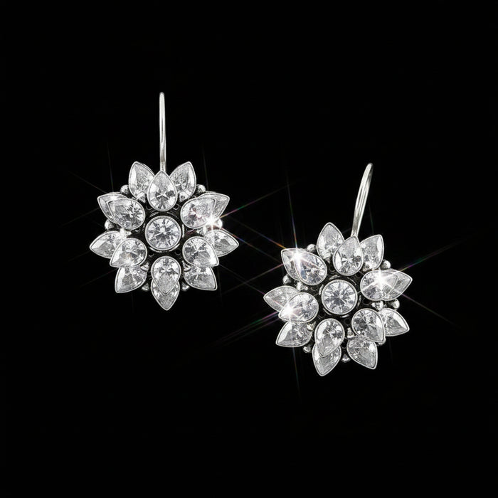 A pair of silver-toned dangle earrings with clear teardrop and round crystals arranged in a floral burst pattern, displayed on a reflective dark gray surface with the brand name SAHELI MY BEST FRIEND at the top.