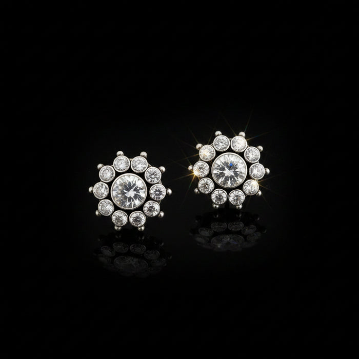 A pair of silver-toned stud earrings with clear round-cut gemstones arranged in a floral or snowflake-like cluster, displayed on a dark reflective surface with the SAHELI MY BEST FRIEND logo above.