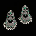 A pair of ornate silver-toned Indian jhumka earrings with green stones and white pearl-like bead drops, displayed on a dark reflective surface. The brand logo 'SAHELI BY SAHELI BRAND' is visible at the top center.