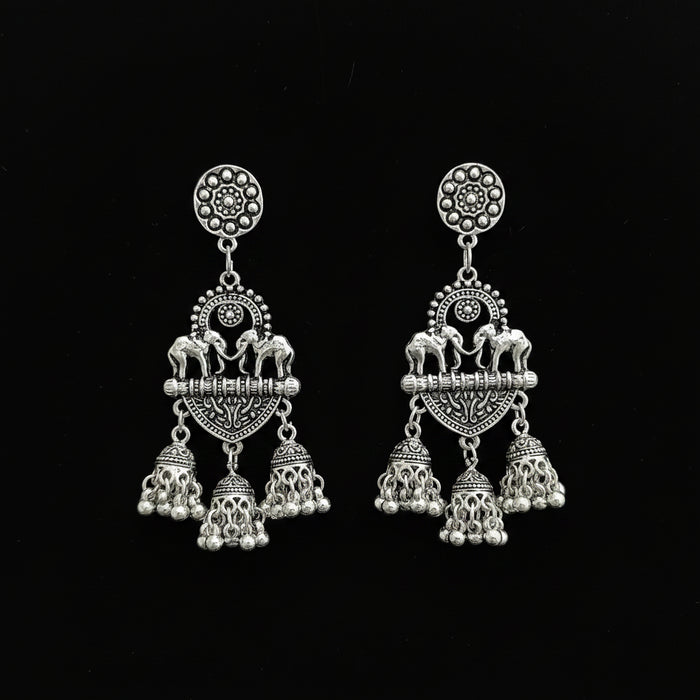 A pair of silver-toned Indian-style dangle earrings with elephant and floral motifs, displayed on a black stand against a dark reflective surface.
