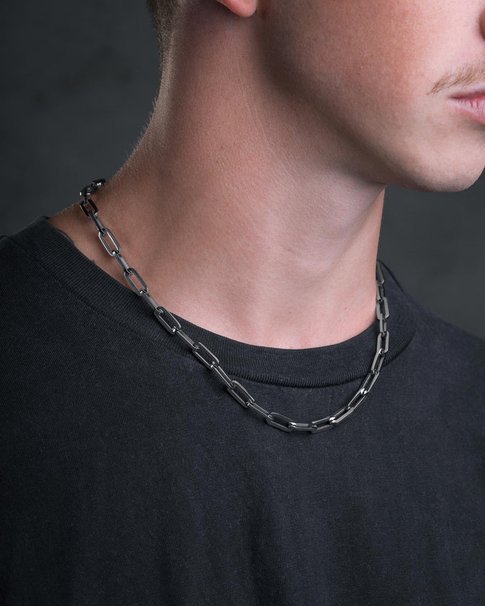 A close-up of a person wearing a black T-shirt and the Statement Collective Paperclip Chain men's necklace, with only the lower part of the face and neck visible.