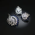 Three floral-shaped jewelry pieces with blue and clear gemstones on a dark background