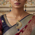 Gold peacock-shaped pendant with blue and red stones on a dark background, branded 'SAHELLI'.