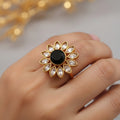 A gold-toned ring shaped like a lotus flower, featuring a central black oval stone surrounded by small pale pink stones, shown on a gray background with a white product tag attached.