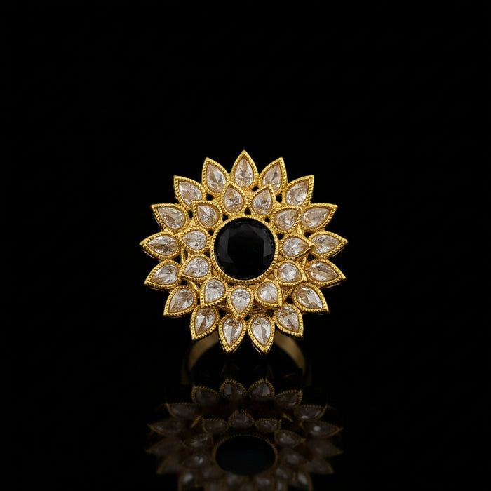 A gold-toned floral brooch featuring a central black oval stone surrounded by clear teardrop-shaped stones, displayed on a dark reflective surface with the SAHELI brand logo in the top left corner.