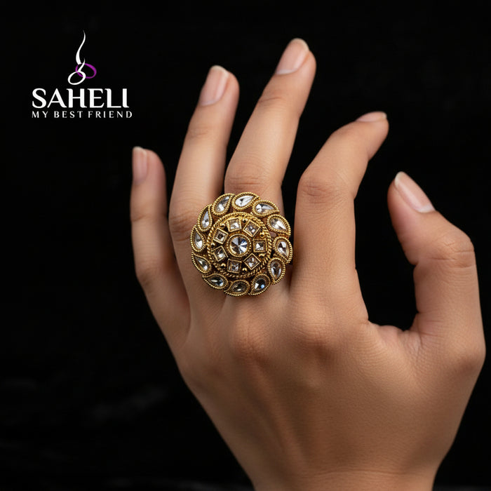 A large, ornate gold ring with intricate stone detailing is displayed on a black velvet glove hand against a dark background. The brand logo 'SAHELI MY BEST FRIEND' is overlaid near the top of the hand.