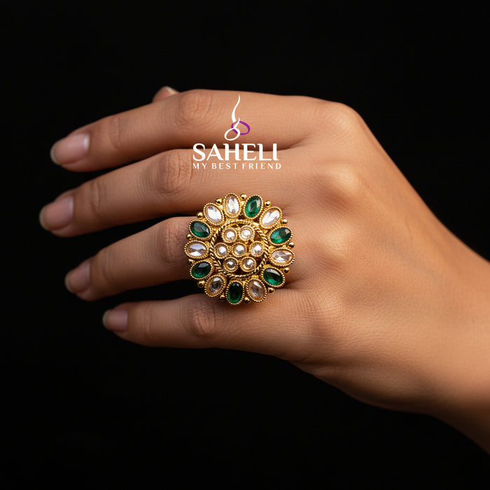 A large gold-toned statement ring with green and clear stones, displayed on a black velvet hand against a black background. The brand logo 'SAHELI MY BEST FRIEND' is visible on the ring.