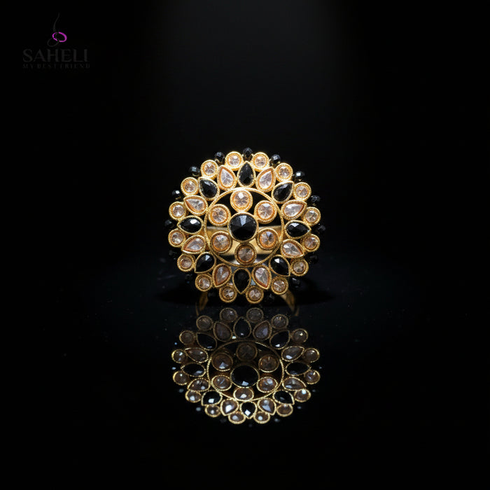 A gold-toned circular Kundan choker brooch featuring black stones and clear stones, displayed on a dark gray reflective surface with a Saheli My Best Friend logo in the top left corner.