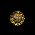 A gold ring with a large, ornate lotus-shaped floral face set with small clear stones, photographed on a dark gray reflective surface. The Saheli brand logo is visible in the top-left corner.