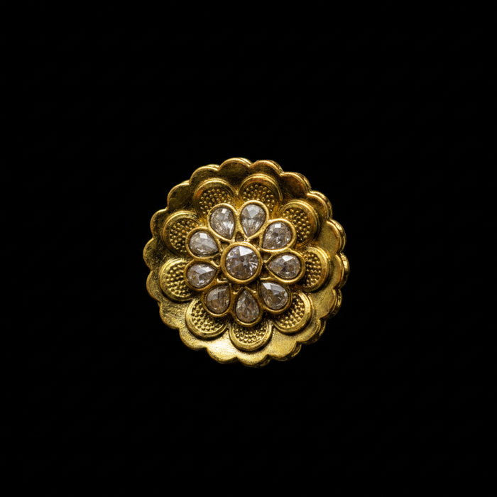 A gold ring with a large, ornate lotus-shaped floral face set with small clear stones, photographed on a dark gray reflective surface. The Saheli brand logo is visible in the top-left corner.