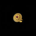 A gold ring with an intricate circular filigree pattern and a small red center stone, displayed on a black hand.