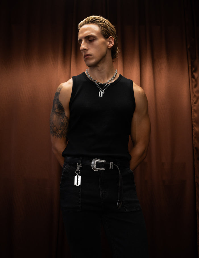 Man wearing black, showcasing a razorblade belt clip standing against a brown curtain.