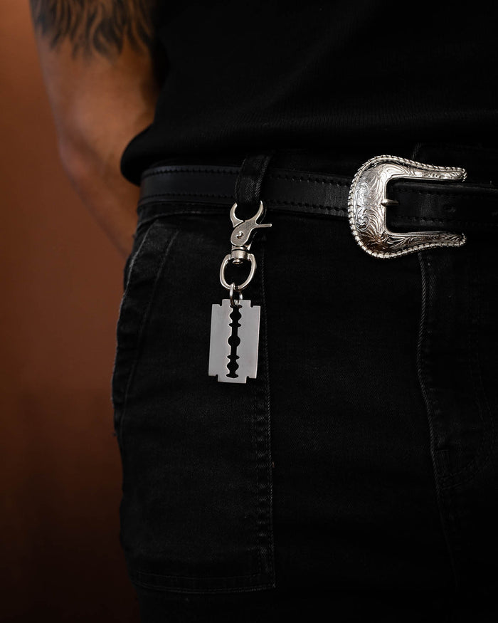 Wearing black jeans, a black shirt, and a silver ornate belt buckle, this person adds an edgy touch to their alternative look with the Razor Blade Belt Clip keychain.
