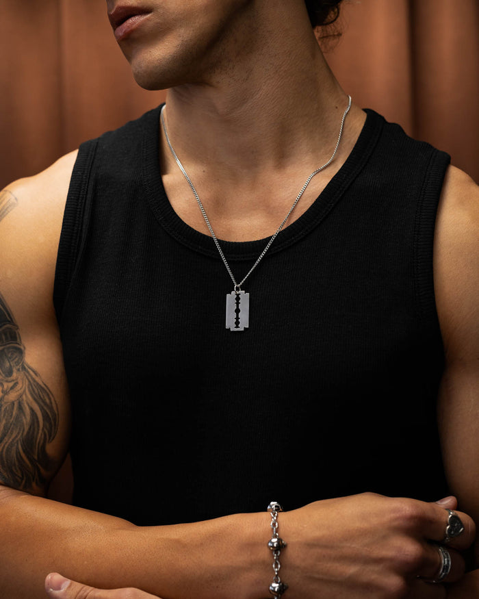 Wearing a black sleeveless shirt, punk bracelet, and a Razor Blade Pendant stainless steel necklace, the tattooed person stands against a brown background.