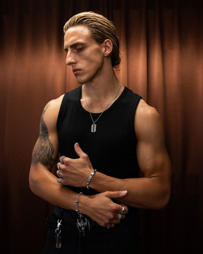 Man wearing a black tank top with tattoos and razorblade necklace against a brown curtain background