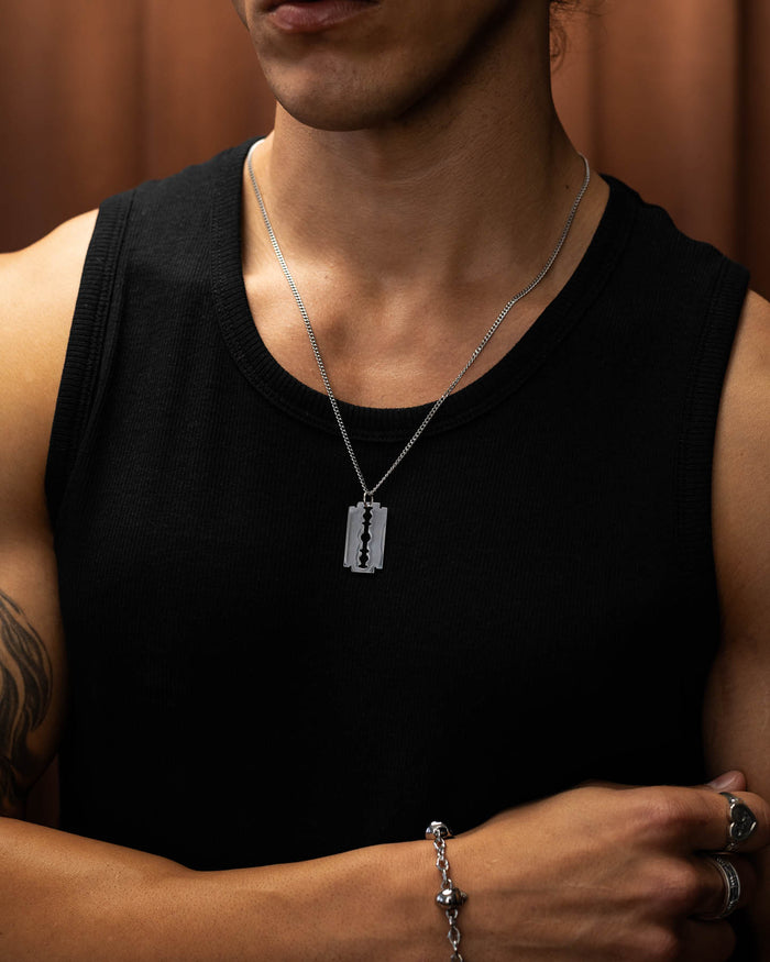 A person wearing a sleeveless black shirt, silver bracelet, and the Razor Blade Pendant stands with one arm crossed. The softly blurred, warm-toned background highlights their edgy punk jewelry look.