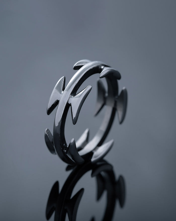 The Razor Wire Ring features pointed arrow-shaped spikes evenly spaced around a stainless steel band, photographed on a reflective dark surface—a bold piece of streetwear jewelry.