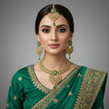 Gold-toned traditional Indian jewelry set featuring a multi-strand necklace, a matching maang tikka, and a pair of chandelier earrings with green stone accents and clear stones, displayed on a dark reflective surface. The brand logo 'SAHeli' is visible at the top center.