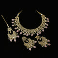 Gold-plated Indian ethnic jewelry set featuring a statement necklace, matching earrings, and a maang tikka, embellished with pink stones and white faux pearls, displayed on a dark reflective surface with the brand name 'SAHELL' visible in the background.