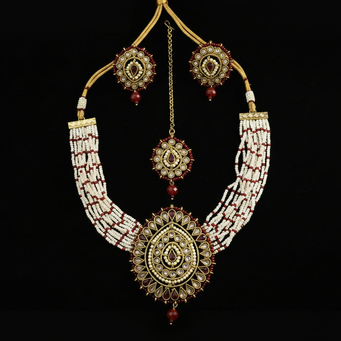 Gold and white beaded traditional Indian jewelry set featuring a necklace, earrings, and a maang tikka, displayed on a dark gray background. The set includes intricate gold-plated details, white seed beads, and red stone accents.