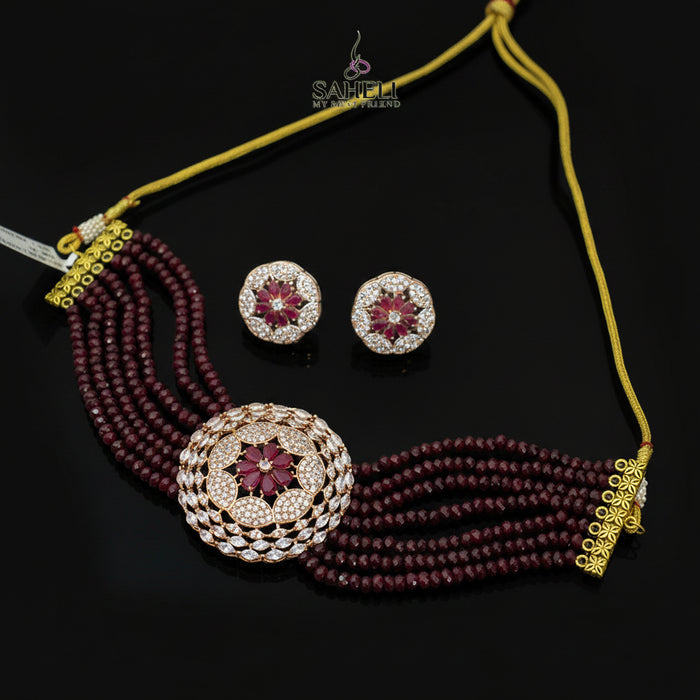 Jewelry set with maroon beads and gold accents on a dark surface, featuring the Saheli brand.