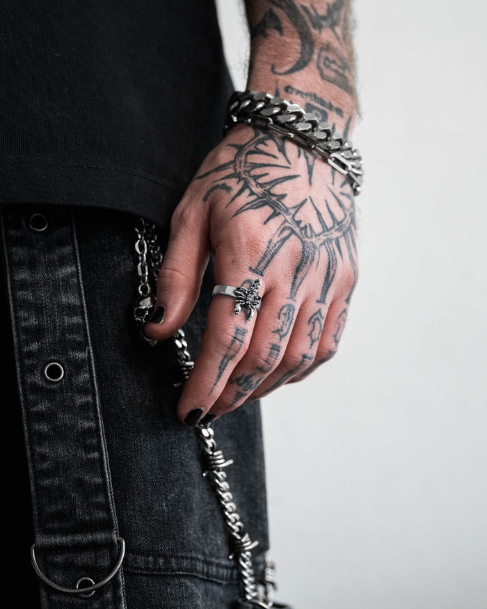 A tattooed hand with black nail polish showcases chunky silver rings, including the Scorpion Ring, and bold bracelets. The wearer’s dark clothing, metal chains, and grommets emphasize an edgy, alternative style.