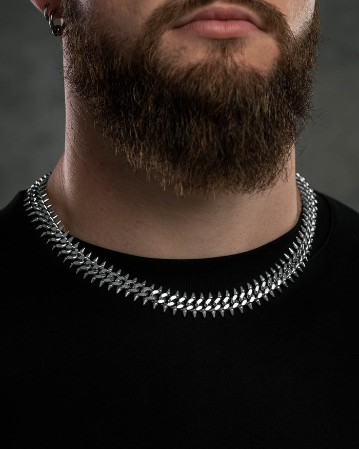 A man with a full brown beard wears a black shirt and a shiny "Screwed Up" Spiked Cuban Chain made from 316L stainless steel. The background is dark and blurred.