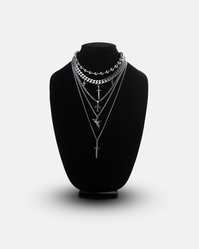 Statement Collective’s Sharp Objects Set is shown on a black velvet necklace display stand, featuring five layered silver chains including a bold Cuban link chain and assorted pendant necklaces with swords, crosses, and geometric accents, all set against a clean white background.