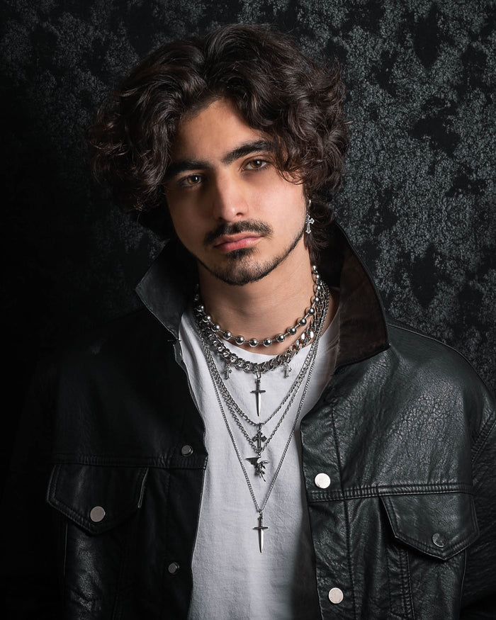A young man with wavy dark hair stands against a dark, textured background, wearing a black leather jacket and white shirt, accessorized with the Statement Collective Sharp Objects Set—layered silver necklaces featuring a cross pendant and a bold Cuban link chain—and silver earrings.