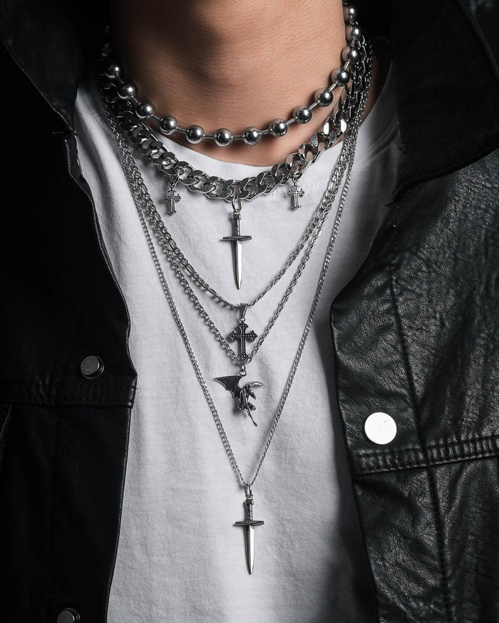 Wearing the Statement Collective Sharp Objects Set, this close-up showcases layered silver necklaces—a bold Cuban link chain and a striking pendant necklace with cross and sword charms—styled over a white T-shirt and accented by a black leather jacket.