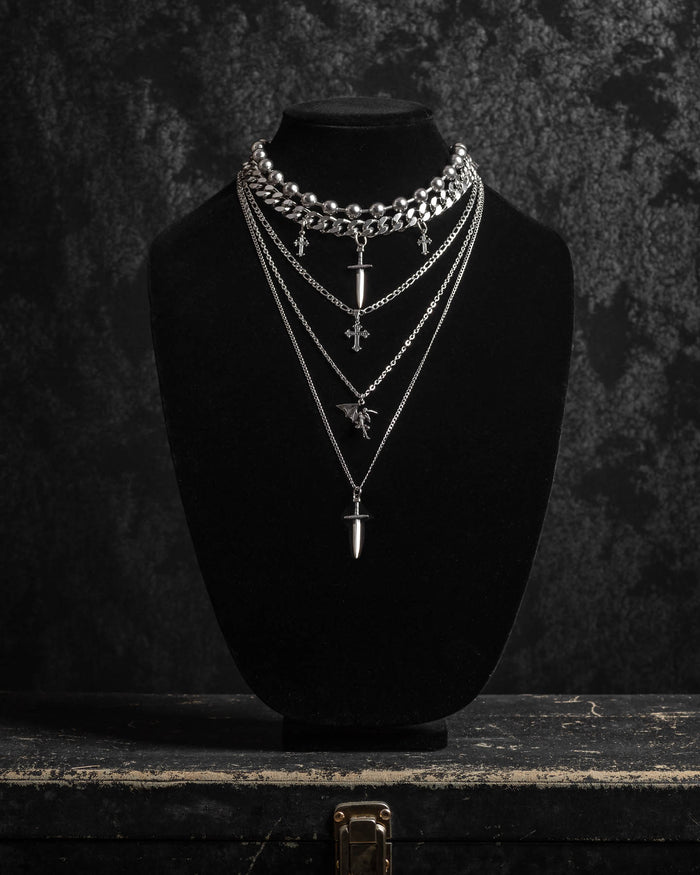 Displayed on a black velvet necklace bust, the Sharp Objects Set by Statement Collective features four layered silver necklaces—including bold Cuban link and striking pendant designs with crosses and bullet-shaped charms—arranged against a dark textured background to create a stunning statement ensemble.