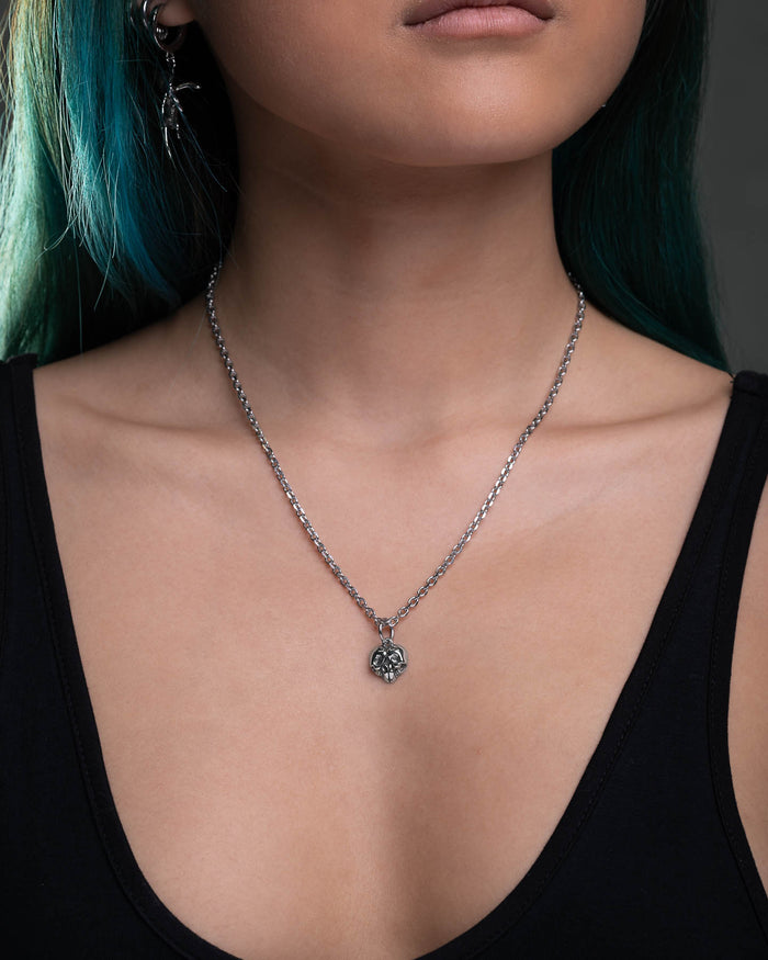 A woman with teal hair models the Skull Heart Pendant—a stainless steel chain with a small ornate skull heart—paired with a black tank top. Only her lower face, neck, and upper chest are visible against a dark background.