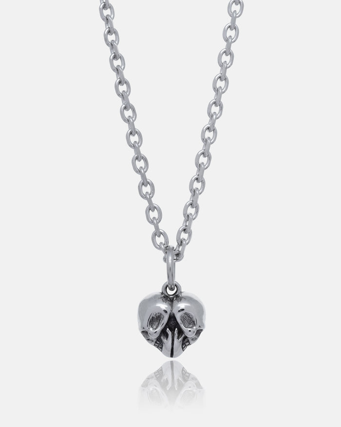 The Skull Heart Pendant is a stainless steel necklace featuring two small skulls joined at the side and facing opposite directions, showcasing dark romance jewelry against a plain white background.