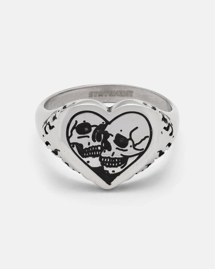 The STATEMENT Skull Heart Ring is a silver heart-shaped ring showcasing two black skulls facing each other engraved on the front, accented with subtle decorative markings along the sides. This bold piece of gothic jewelry is perfect for those who embrace unique and unconventional style.
