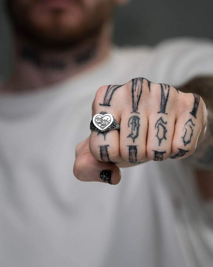 A person in a white shirt holds a fist forward, showcasing the Skull Heart Ring on their finger. The image focuses on the hand and ring, with black arrow tattoos visible and the face blurred, highlighting this unique gothic jewelry piece.