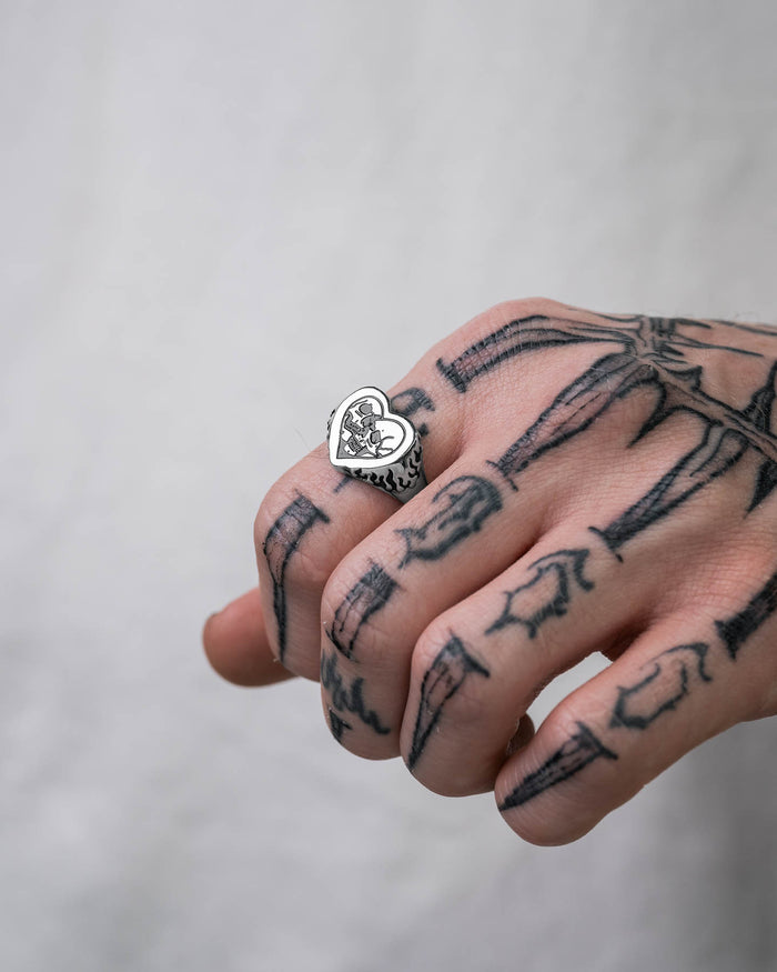 A hand with skeleton tattoos wears the Skull Heart Ring—silver, heart-shaped gothic jewelry with a black illustration—set against a plain, light background.