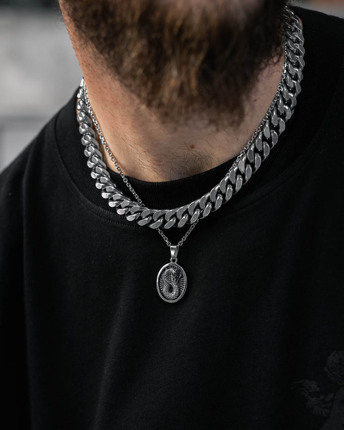 A close-up shows a person wearing two silver chains—a thick, flat one and a thinner chain with the Snake Charmer Pendant, an oval talisman with an engraved design—over a black shirt. The lower face and beard are partially visible.