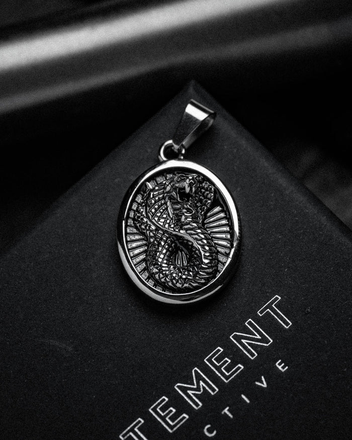 A black and white close-up of the Snake Charmer Pendant, crafted from stainless steel with a detailed dragon design, resting on a black surface where part of the word "TEMENT" is visible.