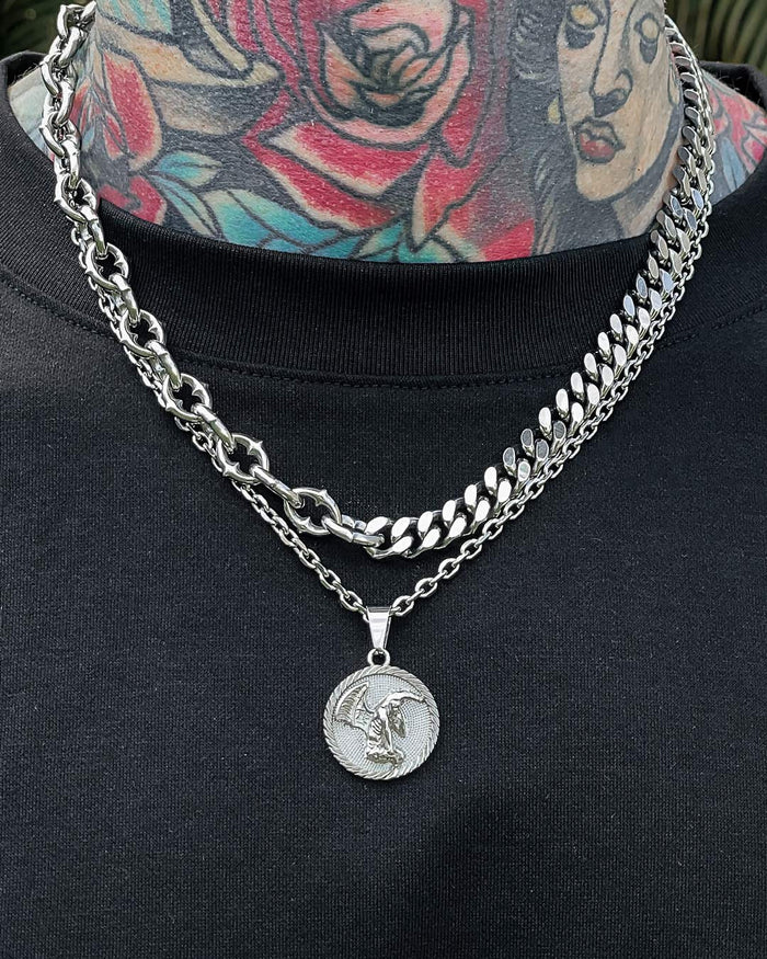 A person with colorful neck tattoos wears gothic jewelry—a spiked chain and the "Split" Cathedral Cuban Chain (Adjustable) with a coin pendant showing a winged figure—over a black shirt. Another chain has a star-shaped design.