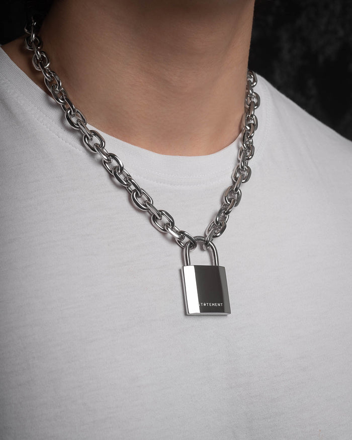 A person wears the Statement Lock & Chain 2.0, a chunky silver necklace with a bold padlock pendant engraved with VETEMENTS, making it the perfect statement jewelry over a plain white t-shirt.