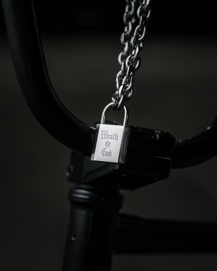 The Statement Lock & Chain 2.0, a thick metal chain with a silver padlock pendant engraved with Wrath of God, is wrapped around black bicycle handlebars against a dark, blurred background.