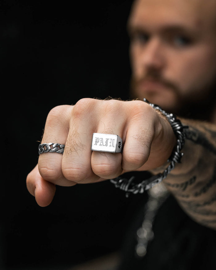 A bearded, tattooed man extends his fist toward the camera, displaying the Statement Signet Ring engraved with "PAIN" on his middle finger, along with silver rings and a chain bracelet. The dark, blurred background heightens the intensity.