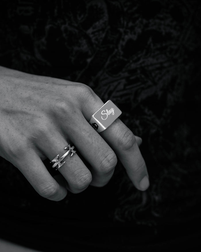 A close-up black-and-white photo shows a hand wearing two rings: the Statement Signet Ring, a chunky stainless steel piece engraved with "Slay," and a thinner crisscross ring, both set against a dark, textured background.